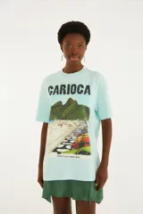 Remera celeste de corte holgado, con cuello redondo, mangas cortas y un gran estampado frontal que dice "CARIOCA" junto a una ilustración de playa y autos.