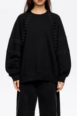 Sudadera negra de corte oversize, confeccionada en algodón, con escote redondo y mangas largas tipo ranglán. Presenta detalles de remaches metálicos distribuidos a lo largo de las costuras de las mangas.