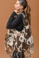Bolso tipo hobo de gran capacidad con estampado animal print de vaca en tonos marrón y blanco. Presenta una etiqueta de cuero con el logo de la marca en el frente y un diseño de caída relajada.
