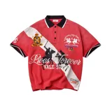 Camiseta polo roja de manga corta con cuello y ribetes en negro y blanco, diseño cruzado con banda diagonal blanca y múltiples bordados y estampados de inspiración equina y deportiva.