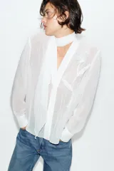 Blusa de tejido semitransparente con efecto arrugado, escote en pico y manga larga. Presenta un detalle decorativo frontal con estructura en forma de flor.