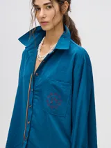 Camisa azul de corte holgado, con cuello camisero, mangas largas con detalles en los puños, bolsillo frontal con bordado y cierre delantero con botones.