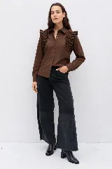 Camisa de color marrón chocolate, con cuello clásico, cierre frontal de botones y mangas largas. Presenta detalles de volados en los hombros que aportan volumen y un estilo distintivo.