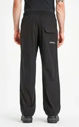 Pantalón negro de tejido técnico impermeable, corte recto y holgado, con elástico en cintura y puños. Presenta cordones ajustables con estampado repetitivo en la bragueta y un pequeño parche cuadrado con un logo de tijeras en el muslo derecho.