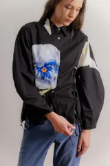 Camisa negra de popelina con diseño de flor azul estampada en el frente. Presenta cuello clásico, mangas abullonadas y detalle de cordones laterales tipo corset para ajustar la silueta.