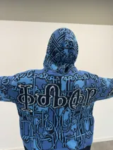 Buzo tipo hoodie tejido de punto grueso con cierre frontal completo hasta la capucha. Presenta un diseño gráfico abstracto en tonos azules y negros, con tipografía estilo gótico en el frente y las mangas. Cuenta con puños y ruedo acanalados en color negro, y etiqueta de marca en la manga.