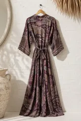 Vestido largo de estilo bata y corte cruzado, con mangas amplias y un lazo ajustable en la cintura. Presenta un estampado de diseño paisley en tonos morados, marrones y azules.