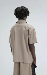 Camisa de manga corta color beige, estilo técnico, confeccionada en microfibra liviana. Cuenta con doble bolsillo frontal, uno con detalle de malla y otro con cierre, y broches en lugar de botones.