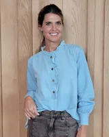 Blusa de denim celeste con cuello alto tipo mao con volados, cierre frontal parcial con botones y mangas largas con puños ajustados.