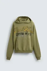 Sudadera con capucha de corte relajado en color verde oliva con efecto lavado. Presenta un bolsillo frontal tipo canguro y un estampado gráfico en el pecho con letras en tonos amarillos y negros. Cuenta con puños y bajo acanalados.