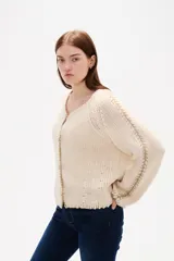Cardigan de punto color natural, con cuello en V, cierre frontal con botones y diseño holgado. Presenta detalles de costuras decorativas bordadas a lo largo de las mangas y en los bordes.