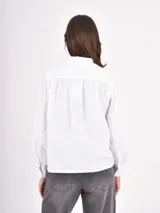 Camisa blanca de manga larga con cuello estilo bebé con volados y puños abotonados. Presenta un cierre frontal con botones a tono.