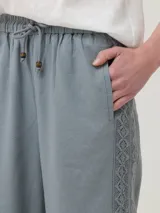 Pantalón de lino orgánico y viscosa color celeste, de corte holgado y tiro alto. Presenta cintura elástica con cordón de ajuste y detalles de encaje broderie tono sobre tono en los laterales y en la parte inferior de las piernas.