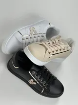 Zapatillas deportivas de cuero sintético en color blanco, beige y negro, con cordones y aplique decorativo dorado con forma de moño.
