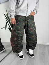 Pantalón cargo baggy con estampa de camuflaje en tonos verdes y marrones, de corte holgado, tiro medio y diseño urbano. Cuenta con detalles de costuras y bordados en hilo rojo a los lados de la pierna.