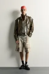 Cazadora cropped fit de color verde militar, confeccionada en tejido efecto piel. Presenta cuello subido, manga larga con puño abotonado, trabillas en los hombros, y bolsillos delanteros de plastrón con solapa. Su diseño incluye bajo elástico y cierre frontal con cremallera oculta por solapa con botones a presión.