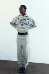 Pantalón jogger gris melange de tiro medio con cintura elástica y puños elásticos en los tobillos. Presenta bolsillos laterales ocultos y un estampado frontal con la palabra "LOS ANGELES" en azul marino.