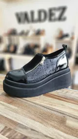 Zapatos de plataforma negros con diseño urbano, confeccionados en una combinación de cuero charol, gamuza y detalles con textura símil cocodrilo. Cuenta con cierre lateral y suela gruesa tipo plataforma.