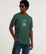Remera de algodón color verde oscuro, con cuello redondo y manga corta. Presenta una estampa frontal en color blanco con el texto 'The Resort Club' y una ilustración de una silla de playa y el sol.
