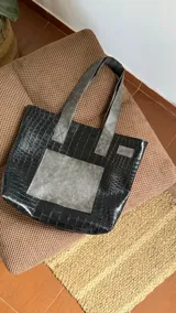 Tote bag de diseño rectangular confeccionada en ecocuero negro con textura de reptil. Presenta un bolsillo frontal cuadrado y correas de hombro en un tono gris desgastado, ofreciendo un contraste moderno y funcional.