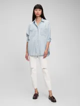 Camisa oversize de jean celeste claro, con cuello clásico, cierre frontal con botones a presión, dos bolsillos con solapa en el pecho y mangas largas con puños abotonados.