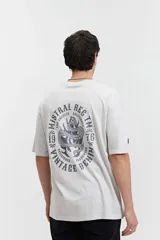 Remera oversize de algodón con efecto acid wash, color blanco hueso. Estampado grande en la espalda con un diseño circular que incluye una serpiente enroscada, texto "MISTRAL REG", "LOS ANGELES", "CALIFORNIA", "VINTAGE DENIM", "REGISTERED" y "TM 76".
