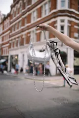 Cartera shoulder bag de cuero metalizado plateado, con diseño tipo banana, asa corta de hombro y correa larga de cadena metálica entrelazada desmontable. Incluye cierre superior.
