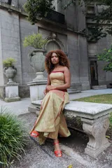 Vestido largo de satén en tono dorado con acabado brillante, presenta un corset strapless con detalles en color bordó en el escote y la cintura, y una falda amplia con costuras decorativas en contraste.