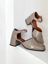 Zapato de cuero vacuno color gris topo, con talón descubierto y pulsera con hebilla. Tiene taco de madera forrado en cuero y suela de goma. La horma es de punta cuadrada, ancha y cómoda.