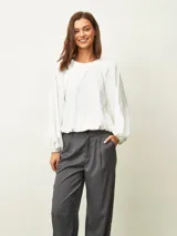 Blusa de color blanco hueso con cuello redondo, mangas largas abullonadas y puños elásticos. Presenta un diseño con frunces en los hombros y ruedo elástico que le otorga una caída holgada.