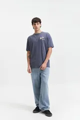 Remera de manga corta color crema, con escote redondo y ajuste regular. Presenta un estampado en la espalda con flores azules y texto en negro que dice "FEEL GOOD" en vertical, "ALL DAY EVERY DAY THE GOOD VIBES CREW mistral" y "ALWAYS IN GOOD COMPANY SINCE 1976".