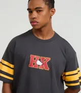 Remera gris oscuro de algodón con corte boxy, cuello redondo y mangas cortas con rayas amarillas. Estampado trasero de Hello Kitty jugando al baloncesto con letras rojas.