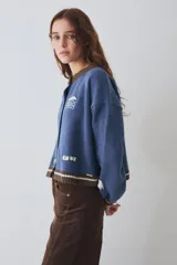 Cardigan de lana color crema con cuello redondo, cierre frontal de botones y mangas largas con puños ajustados. Presenta bordados decorativos en el frente que incluyen un sol, olas y estrellas, con detalles en azul en el cuello y el ruedo.