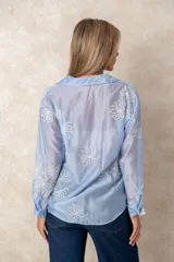 Camisa de manga larga en color blanco, confeccionada en tejido traslúcido con delicados bordados florales en hilo de seda al tono. Presenta cuello clásico, cierre frontal con botones y puños abotonados.