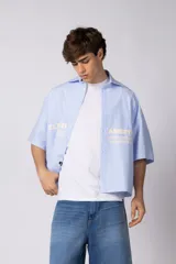 Camisa de manga corta de corte boxy, confeccionada en popelina a rayas celestes y blancas. Presenta cuello camisero clásico, cierre frontal con botones y estampado tipográfico en el pecho.