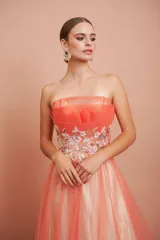 Vestido largo de fiesta, strapless, con busto drapeado y falda de tul con degradé de color coral a blanco. Presenta bordados florales en el corpiño.