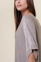 Maxi sweater tejido color beige con efecto metalizado sutil, silueta amplia, mangas tipo murciélago y largo asimétrico (más corto adelante y más largo atrás).