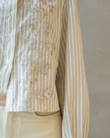 Camisa de corte clásico a rayas verticales en tonos neutros, con cuello camisero y cierre frontal de botones. Presenta un delicado bordado floral con canutillos y lentejuelas brillantes en el frente.