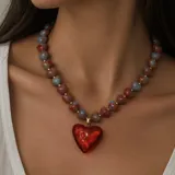 Collar corto compuesto por cuentas acanaladas en forma de corazón en tonos grisáceos y rojizos con pequeñas cuentas doradas intermedias, del cual cuelga un gran dije central en forma de corazón rojo translúcido con detalles dorados.