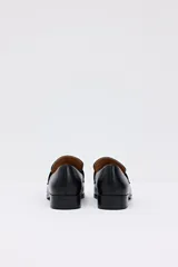 Mocasín de cuero negro con detalle de hebilla metálica plateada en el empeine y suela plana.
