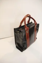 Cartera tipo bowling color beige con detalles en cuero negro, doble asa corta y cierre superior con cremallera.