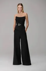 Mono largo strapless color negro, con diseño asimétrico en el torso, bolsillos laterales, pantalón amplio de corte recto y cinturón incorporado con hebilla metálica.