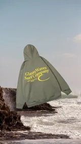 Hoodie de corte oversize color verde militar, con capucha y bolsillo tipo canguro. Presenta un estampado gráfico en la espalda con el texto 'Glassy Waves Surf Company' y un logotipo de ola en tono amarillo.