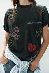 Camiseta oversize de algodón con efecto lavado, cuello redondo y manga corta. Presenta múltiples aplicaciones de brillos y strass con motivos coloridos de Smiley, incluyendo estrellas, corazones y caras sonrientes.