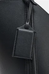 Bolso tipo shopper de hombro en color beige, con diseño estructurado, doble asa y detalle de pespuntes verticales en el cuerpo. Incluye un colgante decorativo tipo etiqueta y cierre imantado.