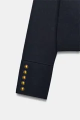 Chaqueta de punto de estilo militar, color azul marino, con cuello redondo y manga larga. Presenta cierre frontal con corchetes ocultos y detalle de botones dorados decorativos en el frente y en los puños.