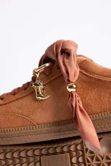 Zapatillas de gamuza color marrón con suela de goma color caramelo. Presentan cordones al tono y un detalle decorativo de pañuelo estampado atado en los laterales con esferas metálicas doradas.