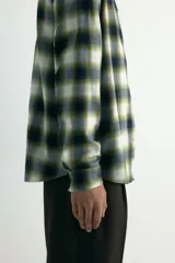 Camisa de franela de algodón suave con cuello inglés, cierre frontal de botones, mangas largas con puños abotonados y bajo redondeado. Presenta un diseño a cuadros en tonos oscuros y amarillentos.