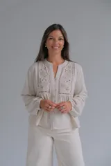 Blusa de color beige con cuello mao y cierre frontal de botones. Presenta detalles de bordado floral y calados en el frente, con mangas largas de corte holgado.