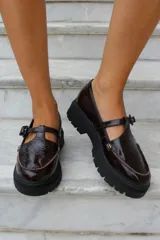 Zapato tipo guillermina en cuero charol color bordó, con suela track negra de goma, cierre con hebilla ajustable y detalle de herrajes metálicos dorados.